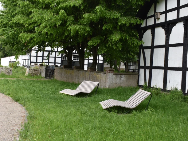 Hille Reimlers Teich Zwei Holzliegen stehen auf einer Wiese unter einem großen Baum neben einem Fachwerkhaus.