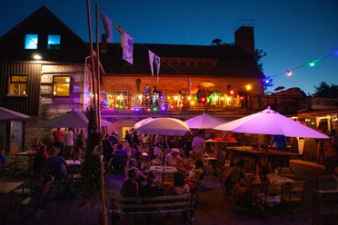 KulturOrt Heymannbaude Ein lebhaftes Abendfest im Freien mit bunten Lichtern, Menschen unter Sonnenschirmen und einer Terrasse mit Musikern vor einem beleuchteten Gebäude.