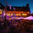 KulturOrt Heymannbaude Ein lebhaftes Abendfest im Freien mit bunten Lichtern, Menschen unter Sonnenschirmen und einer Terrasse mit Musikern vor einem beleuchteten Gebäude.A lively outdoor evening party with colorful lights, people under umbrellas and a terrace with musicians in front of an illuminated building.Živá večerní párty pod širým nebem s barevnými světly, lidmi pod slunečníky a terasou s hudebníky před osvětlenou budovou.Tętniąca życiem wieczorna impreza na świeżym powietrzu z kolorowymi światłami, ludźmi pod parasolami i tarasem z muzykami przed oświetlonym budynkiem.Een levendig avondfeest in de buitenlucht met kleurrijke lichtjes, mensen onder parasols en een terras met muzikanten voor een verlicht gebouw.Una vivace festa serale all'aperto con luci colorate, persone sotto gli ombrelloni e una terrazza con musicisti davanti a un edificio illuminato.