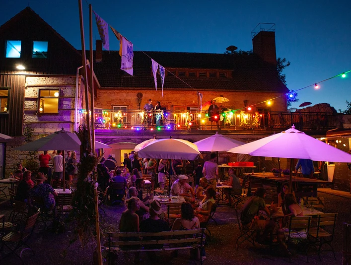 KulturOrt Heymannbaude Ein lebhaftes Abendfest im Freien mit bunten Lichtern, Menschen unter Sonnenschirmen und einer Terrasse mit Musikern vor einem beleuchteten Gebäude.