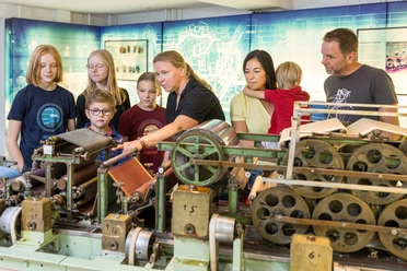 Familie an der Laborpapiermaschine Eine Gruppe von Erwachsenen und Kindern betrachtet eine historische Webmaschine in einem Museum.