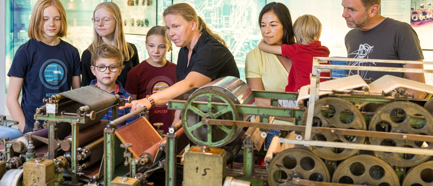 Familie an der Laborpapiermaschine Eine Gruppe von Erwachsenen und Kindern betrachtet eine historische Webmaschine in einem Museum.