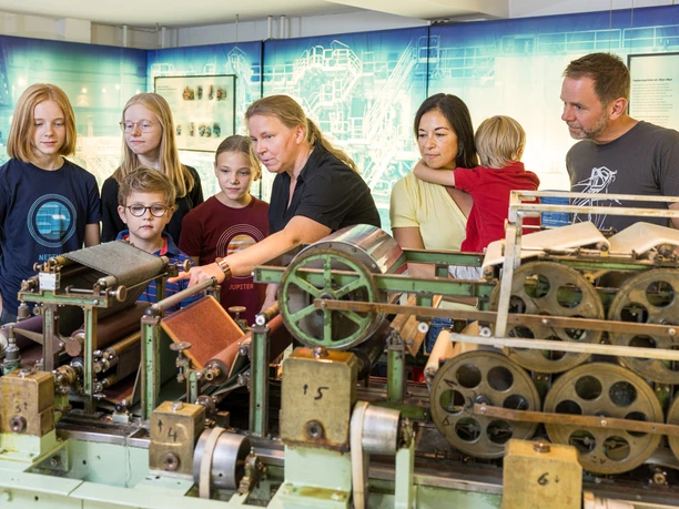 Familie an der Laborpapiermaschine Eine Gruppe von Erwachsenen und Kindern betrachtet eine historische Webmaschine in einem Museum.