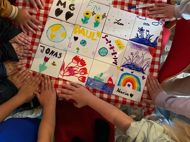 ELML_FlotteFliesen.jpg Kinder gestalten bunte Fliesen mit Namen und Motiven auf rotem Tisch, Hände liegen drumherum.