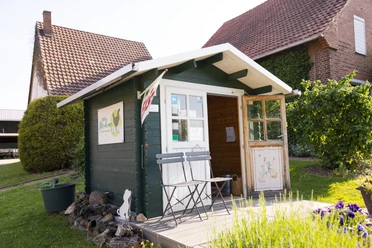 Biohof Mohme_Hofladen Kleiner Hofladen aus Holz mit Vordach, davor zwei Holzbänke; ländliches Umfeld, Sonnenschein.