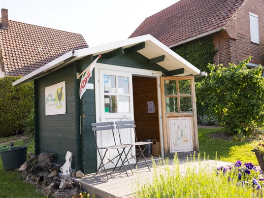 Biohof Mohme_Hofladen Kleiner Hofladen aus Holz mit Vordach, davor zwei Holzbänke; ländliches Umfeld, Sonnenschein.
