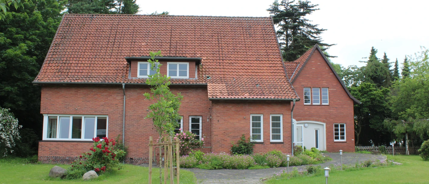Binderhaus Rodewald Ein rotes Backsteinhaus mit Satteldach steht inmitten einer grünen Wiese, umgeben von hohen Bäumen.