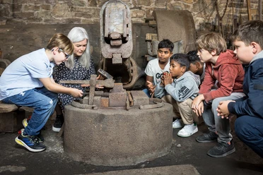 LVR-Industriemuseum Oelchenshammer, Kinder versammelt um eine ältere Person, die Werkzeuge in einer historischen Schmiede erklärt.