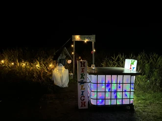 Lichterabend_im Maislabyrinth_IMG_3659.JPG Beleuchteter kleiner Stand mit einem Schild 'HERZLICH' und bunten Lichtern vor einem dunklen Feld mit Maispflanzen