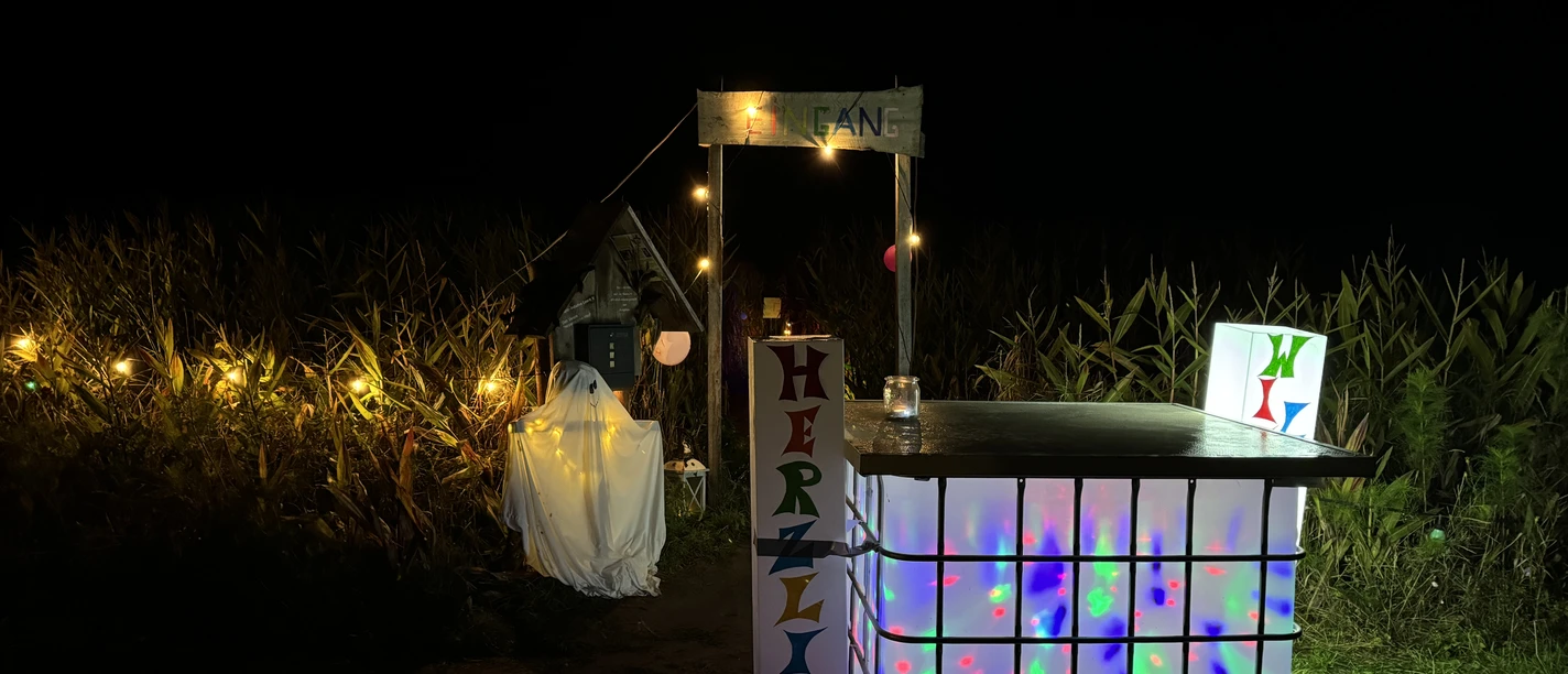 Lichterabend_im Maislabyrinth_IMG_3659.JPG Beleuchteter kleiner Stand mit einem Schild 'HERZLICH' und bunten Lichtern vor einem dunklen Feld mit Maispflanzen