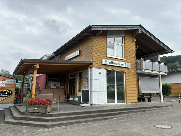 Zweistöckiges Holzgebäude mit Biergarten, Firmenschild "Im Himmelchen", umgeben von Natur.