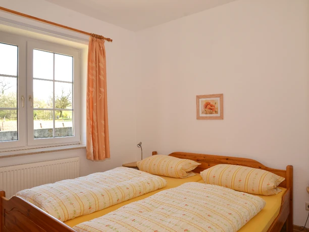 Schlafzimmer Hühnerhof.jpg Helles Schlafzimmer mit Holzdoppelbett, gelber Bettwäsche und Fenster mit Blick in die Natur.