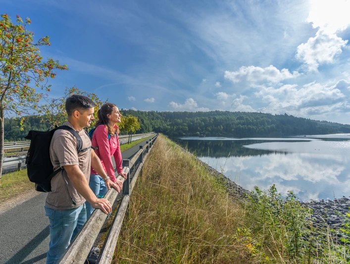 Zwei Personen genießen die ruhige Aussicht auf einen großen Stausee mit umliegendem Waldgebiet.