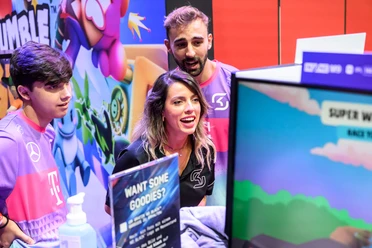 gamescom Eine dynamische Szene bei der Gamescom: Drei Personen in lila T-Shirts interagieren begeistert mit einem Videospiel auf einem Bildschirm.