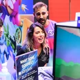 gamescom Eine dynamische Szene bei der Gamescom: Drei Personen in lila T-Shirts interagieren begeistert mit einem Videospiel auf einem Bildschirm.A dynamic scene at Gamescom: three people in purple T-shirts interact enthusiastically with a video game on a screen.