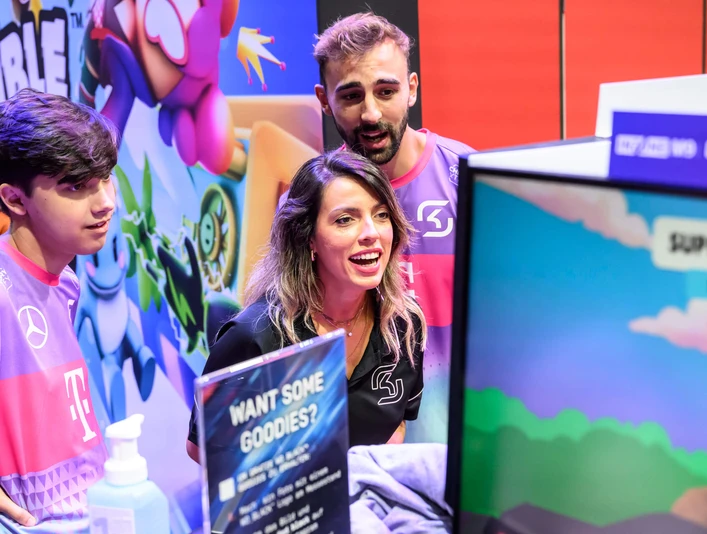 gamescom Eine dynamische Szene bei der Gamescom: Drei Personen in lila T-Shirts interagieren begeistert mit einem Videospiel auf einem Bildschirm.A dynamic scene at Gamescom: three people in purple T-shirts interact enthusiastically with a video game on a screen.