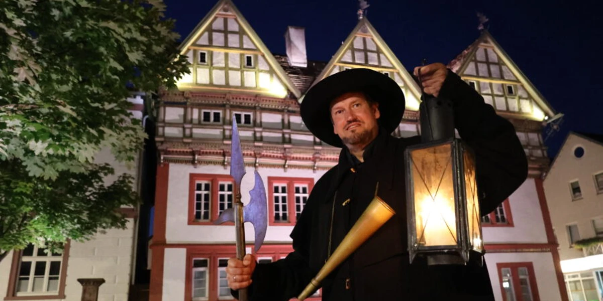 Ein Nachtwächter mit Laterne und Hellebarde steht vor einem beleuchteten Fachwerkhaus bei Nacht.