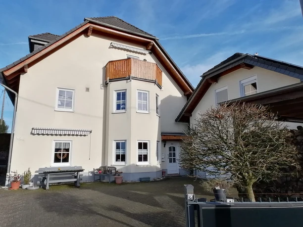 2024_11Haus (1).jpg Großes, modernes Einfamilienhaus mit hellem Putz, schrägem Dach und Balkon, umgeben von Hofpflanzen.