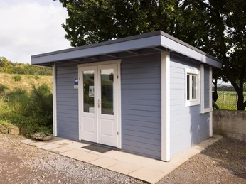 Biohof Wiehenglück_Hofladen Ein blau gestrichenes Holzgebäude mit weißen Türen und Fenstern steht auf einer gepflasterten Fläche.