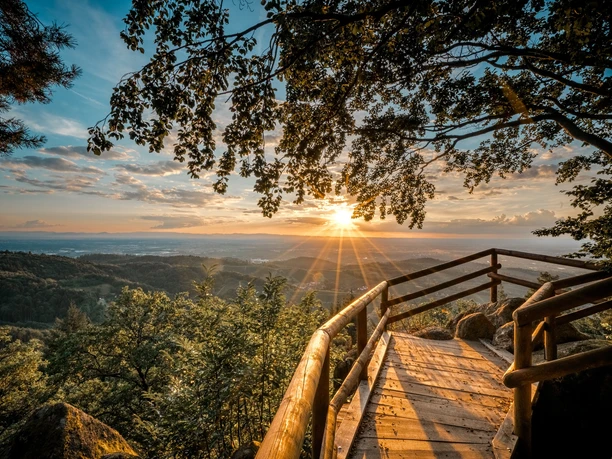 Bürstenstein. Eine Holzplattform unter einem Baum. Man schaut über Felsen und Berge in den Sonnenuntergang