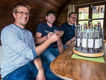 Drei Männer mit Brille, alle mit Jeans und T-Shirt sitzen an einem Tisch mit Schnapsflaschen und prosten einander zu