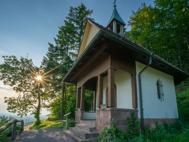 Blosenkopfkapelle Kappelrodeck mit einem Portal aus rotem Sandstein und einem kleinen Glockenturm. Im Hintergrund scheint die Sonne durch eine Baumkrone