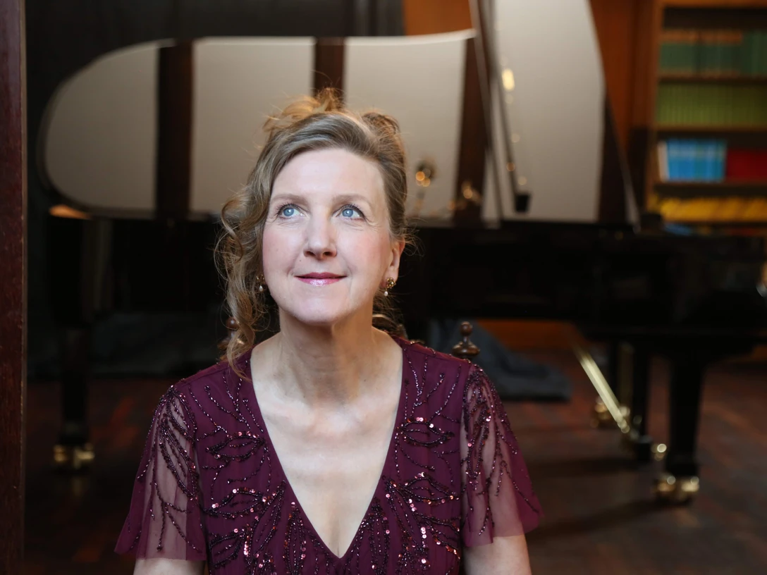 Lohmer März 2024.JPG Eine Frau in einem eleganten, rot schimmernden Kleid sitzt vor einem Flügel und schaut mit einem leichten Lächeln die Decke an. Der Raum ist mit Bücherregalen und einem warmen Holzinterieur ausgestattet.