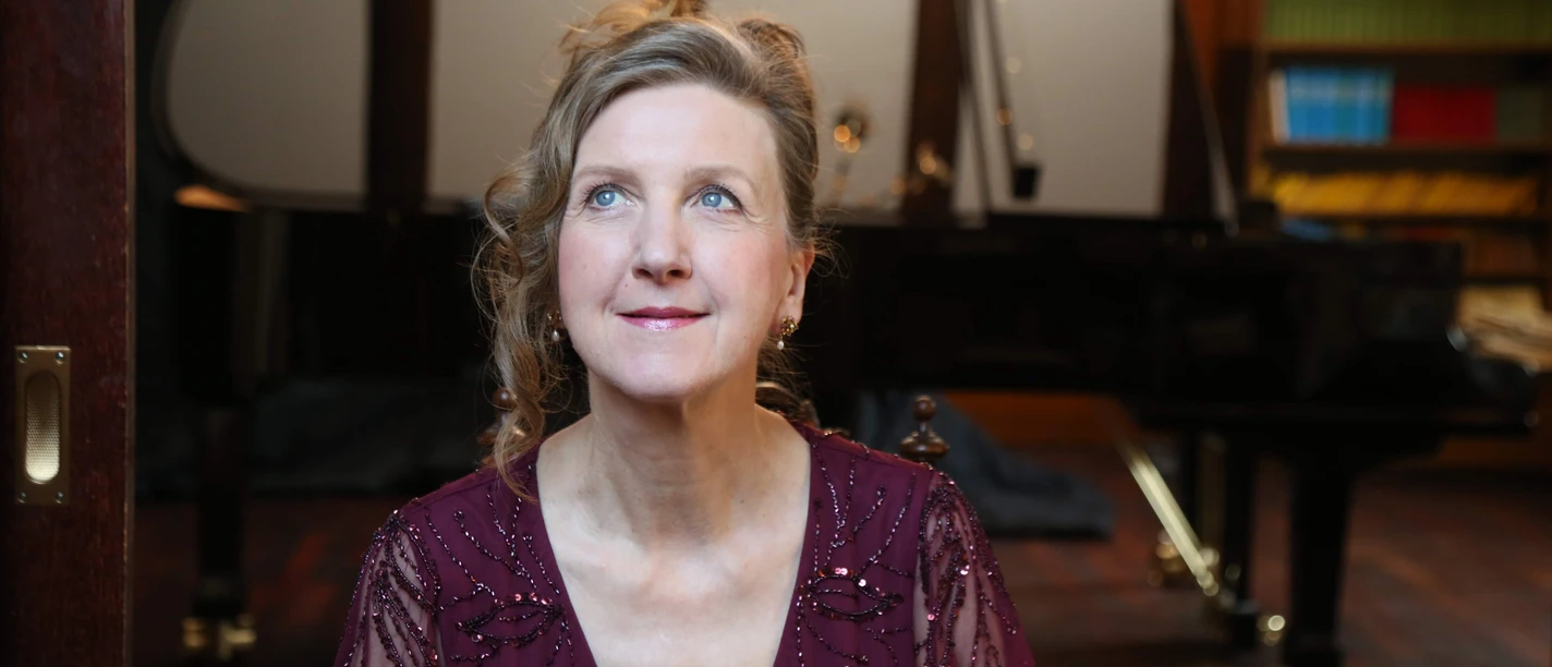 Lohmer März 2024.JPG Eine Frau in einem eleganten, rot schimmernden Kleid sitzt vor einem Flügel und schaut mit einem leichten Lächeln die Decke an. Der Raum ist mit Bücherregalen und einem warmen Holzinterieur ausgestattet.