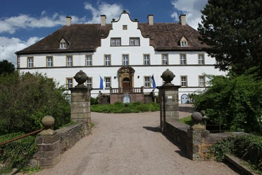 Historisches Schlossgebäude mit weißer Fassade und prächtigem Eingang, umgeben von grünem Garten.