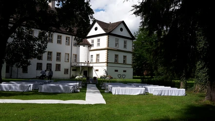 Weiß gedeckte Tische und Stühle stehen auf einer grünen Wiese vor einem historischen Schlossgebäude.