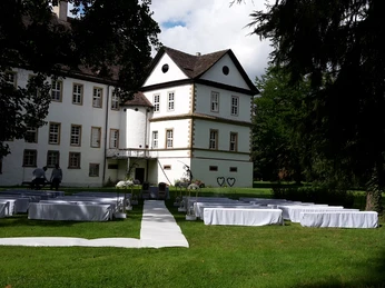 Weiß gedeckte Tische und Stühle stehen auf einer grünen Wiese vor einem historischen Schlossgebäude.