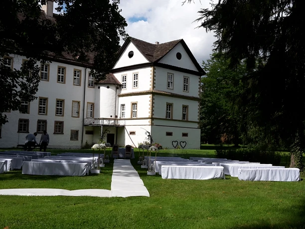 Trauung im Schlosspark Wehrden Weiß gedeckte Tische und Stühle stehen auf einer grünen Wiese vor einem historischen Schlossgebäude.