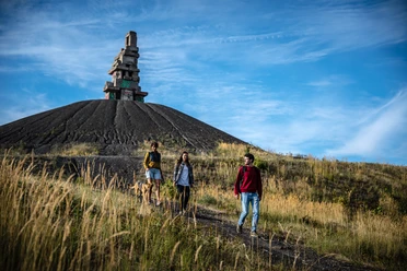 Drei Personen wandern an einem grasbewachsenen Hügel mit einem imposanten Steinmonument oben.