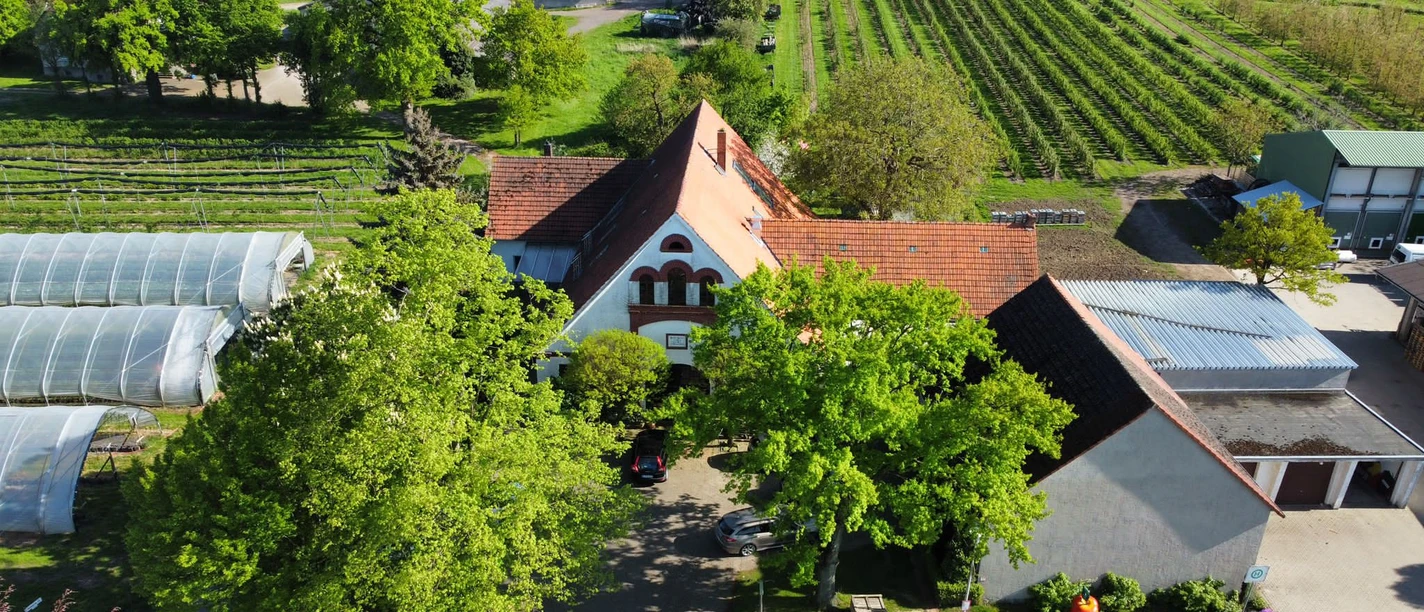 Obsthof Wickemeyer Luftaufnahme des Obsthofs mit Gebäude, umgeben von Gärten und Gewächshäusern, bei sonnigem Wetter.