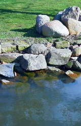 Steine am Wasserufer bei Wunstorf, grüne Wiese im Hintergrund, ruhiges Gewässer im Vordergrund.