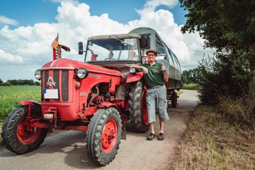 2021-07-25_Planwagen-Sigwardskirche_68-Bearbeitet.jpg Ein fröhlicher Mann steht neben einem roten Traktor mit Planwagen auf einer ländlichen Straße.