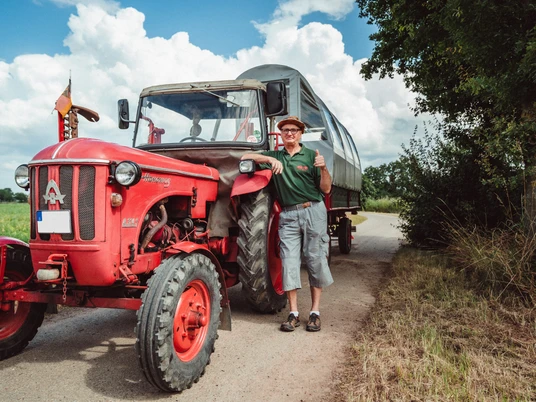 2021-07-25_Planwagen-Sigwardskirche_68-Bearbeitet.jpg Ein fröhlicher Mann steht neben einem roten Traktor mit Planwagen auf einer ländlichen Straße.