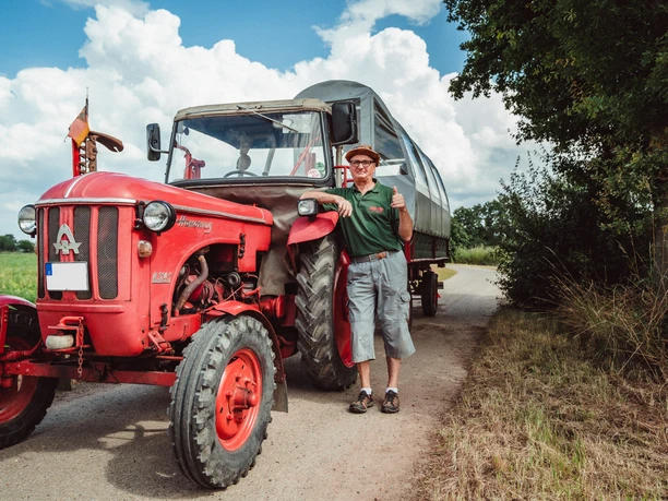2021-07-25_Planwagen-Sigwardskirche_68-Bearbeitet.jpg Ein fröhlicher Mann steht neben einem roten Traktor mit Planwagen auf einer ländlichen Straße.