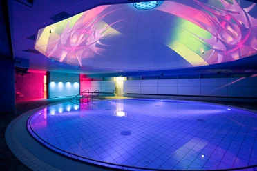 Liquid Sound Tempel Toskana Therme Verlicht binnenzwembad met rond bad, blauwe en roze lichten en kleurrijke projecties op het plafond, moderne en ontspannen sfeer.