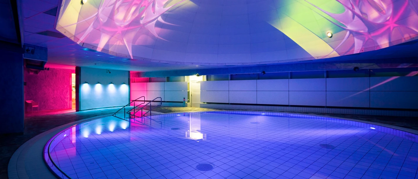 Liquid Sound Tempel Toskana Therme Beleuchtetes Hallenbad mit rundem Becken, blauen und pinken Lichtern sowie bunten Projektionen an der Decke, moderne und entspannte Atmosphäre.