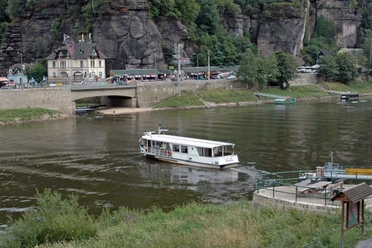 A white ferry sails from Schöna on a river in front of a stone bridge to Hrensko; a building and wooded rocks can be seen in the background.