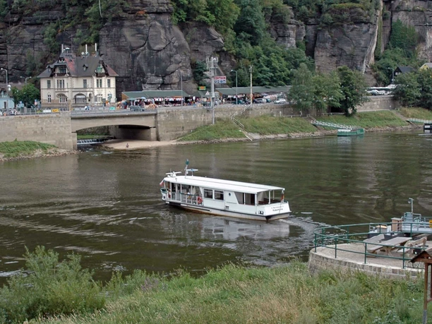 Fähre Schöna Hrensko Eine weiße Fähre fährt von Schöna auf einem Fluss vor einer steinernen Brücke nach Hrensko, im Hintergrund sind ein Gebäude und bewaldete Felsen zu sehen.