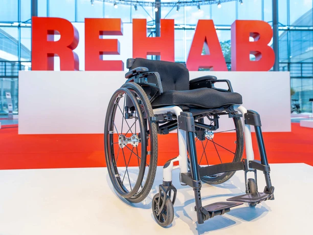 rehab_2023_messekarlsruhe_juergenroesner_165.jpg Rollstuhl vor großem REHAB-Schriftzug in einer modernen Messehalle.