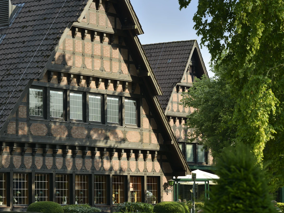 Fassade-09.jpg Backsteinfachwerkhaus mit hohen Giebeln, dunklen Holzbalken und begrüntem Vorgarten bei Sonne.