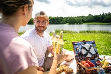 Espelkamp- See am Kleihügel Zwei Personen genießen ein Picknick am See, umgeben von Natur, mit Essen und Getränken im Freien.