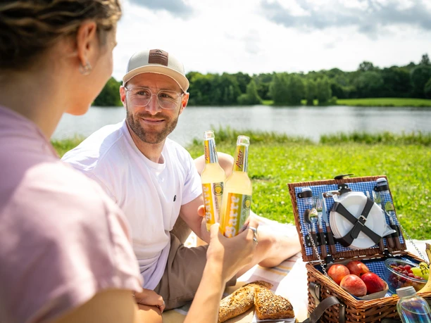 Espelkamp- See am Kleihügel Zwei Personen genießen ein Picknick am See, umgeben von Natur, mit Essen und Getränken im Freien.