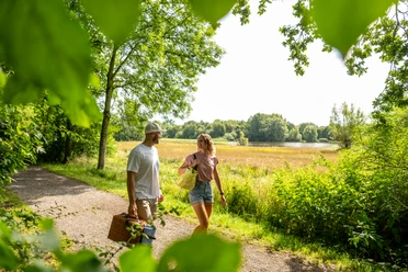 Espelkamp-See am Kleihügel Ein Paar spaziert mit Picknickkorb auf einem Pfad am Espelkamp-See, umgeben von grünen Bäumen.