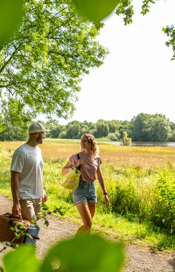 Espelkamp-See am Kleihügel Ein Paar spaziert mit Picknickkorb auf einem Pfad am Espelkamp-See, umgeben von grünen Bäumen.