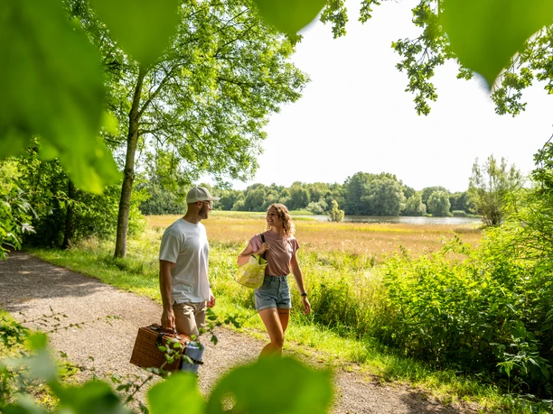 Espelkamp-See am Kleihügel Ein Paar spaziert mit Picknickkorb auf einem Pfad am Espelkamp-See, umgeben von grünen Bäumen.