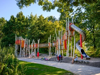 Moderner Abenteuerspielplatz mit Rutschen, Klettergerüsten und Besuchern umgeben von grüner Natur.
